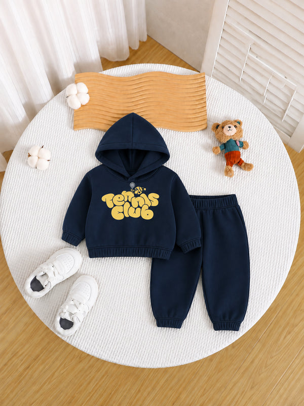 A children's navy blue two-piece outfit set consisting of a hooded sweatshirt with a yellow 'Tennis Club' graphic and matching jogger pants, styled in a flat lay on a round white rug with white sneakers, a teddy bear, and decorative props.